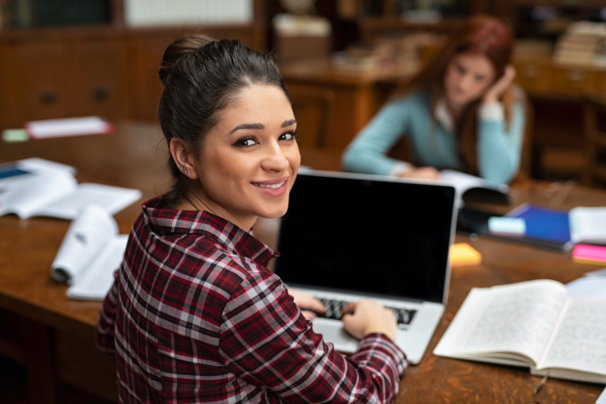Uśmiechnięta studentka siedząca w czytelni z laptopem i książkami